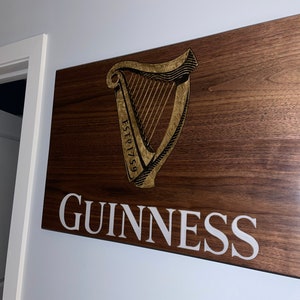 Pu&ograve; includere: Un cartello in legno con il logo Guinness, che presenta un'arpa dorata con il testo "ESTD 1759" e la parola "GUINNESS" in lettere bianche.