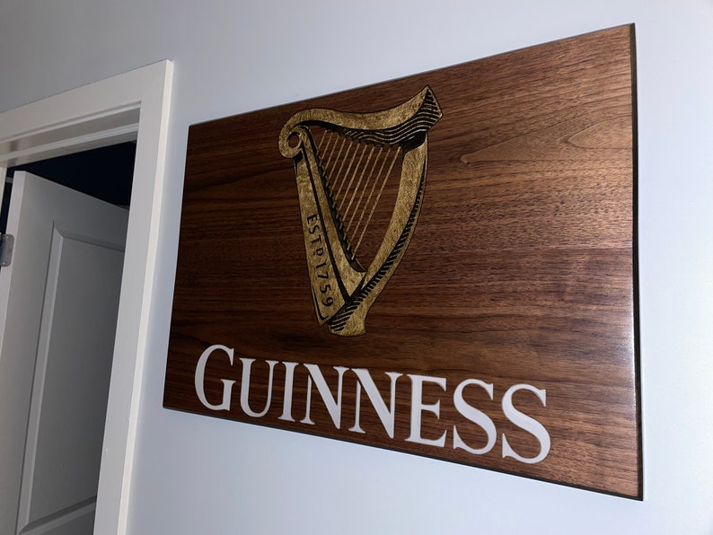 Pu&ograve; includere: Un cartello in legno con il logo Guinness, che presenta un'arpa dorata con il testo "ESTD 1759" e la parola "GUINNESS" in lettere bianche.