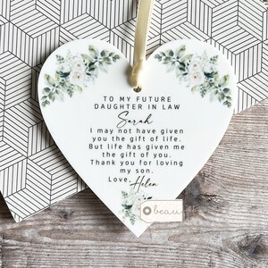 Könnte beinhalten: Weißer herzförmiger Keramik-Anhänger mit Blumenmuster und dem Text "To my future daughter in law Sarah I may not have given you the gift of life. But life has given me the gift of you. Thank you for loving my son. Love, Helen"