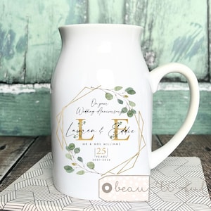 以下が含まれることがあります： 白いセラミックピッチャー、曲がったハンドル付き。金色の幾何学模様と「On your Wedding Anniversary, Lauren & Eddie, 25 Years 2001-2026」の文字が特徴です。デザインには緑の葉のアクセントが含まれています。