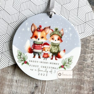 Könnte beinhalten: Ein runder, weißer Weihnachtsbaumschmuck mit einer Schneelandschaft, auf der drei Füchse in Winterkleidung zu sehen sind. Der Text lautet "Daddy - Noah - Mummy First Christmas as a family of 3 2023".