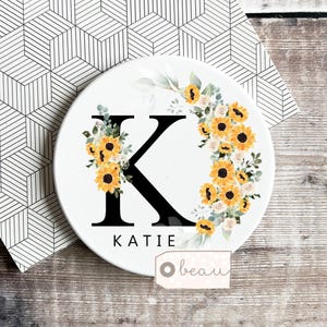 Może przedstawiać: Biały ceramiczny podkładka pod kubek z dużą czarną literą "K" i imieniem "KATIE" poniżej. Litera otoczona jest aranżacją kwiatową z żółtych słoneczników i białych róż. Widoczna jest mała etykieta ze słowem "beau".