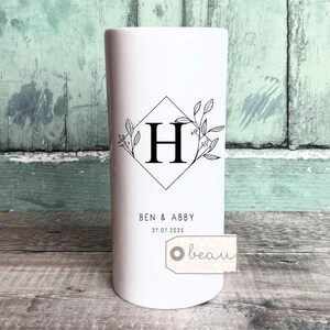 Puede incluir: Jarrón cilíndrico blanco con un diseño en forma de diamante negro que contiene la letra "H" y elementos florales. El texto "BEN & ABBY" y la fecha "27.07.2025" están impresos debajo. Una pequeña etiqueta con la palabra "beau" está adjunta.