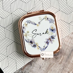 May include: A compact mirror with a rose gold frame and a white background. The mirror features a floral design with a heart shape and the name "Sarah" in cursive writing.