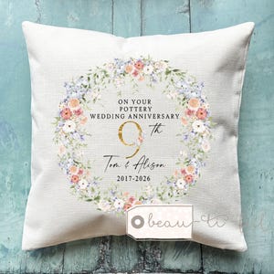 Può includere: Un cuscino quadrato bianco sporco con un design a ghirlanda floreale. La ghirlanda circonda il testo "ON YOUR POTTERY WEDDING ANNIVERSARY 9th Tom & Alison 2017-2026". La ghirlanda presenta fiori rosa, pesca e blu con foglie verdi.