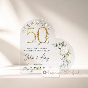 Könnte beinhalten: Eine herzförmige, transparente Acrylplatte mit der Zahl 50 in Goldfolie, weißen Blumenillustrationen und dem Text: 50 years ON YOUR GOLDEN WEDDING ANNIVERSARY John & Amy 1976-2026. Das Design enthält grüne Eukalyptusblätter und ein kleines goldenes Herz.
