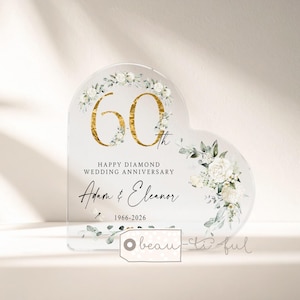 May include: A heart-shaped clear acrylic plaque featuring a gold-colored number 60, white floral illustrations, and the text: Happy Diamond Wedding Anniversary, Adam & Eleanor, 1966-2026. The design includes green leaves and a small dragonfly graphic.