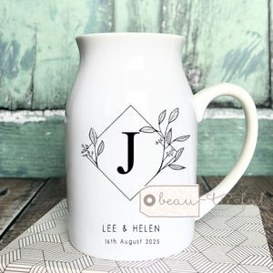 Op de afbeelding: Witte keramische mok met een groot handvat, met een zwarte letter "J" in een ruitvormig frame, versierd met bloemmotieven. De mok heeft de namen "LEE & HELEN" en de datum "16 augustus 2025" erop gedrukt.