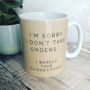 Op de afbeelding: Witte keramische mok met een beige achtergrondpatroon van witte pijlen. De mok heeft zwarte tekst die luidt: "Het spijt me, ik neem geen bestellingen aan... Ik neem nauwelijks suggesties aan."