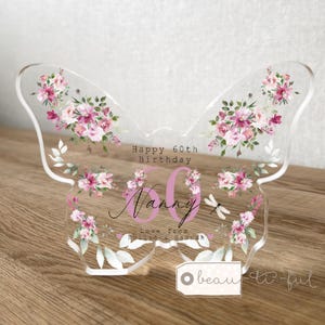 Può includere: Una targa in acrilico trasparente a forma di farfalla con illustrazioni floreali rosa e viola, foglie verdi e una piccola libellula. Il testo recita Happy 60th Birthday, Nanny, Love from Hollie & Hannah, con un grande numero 60 rosa al centro.