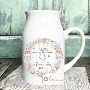 May include: White ceramic pitcher with a handle, featuring a floral wreath design. The text reads "ON YOUR POTTERY WEDDING ANNIVERSARY 9th Grace & Harry 2017-2026". A tag with the word "beau" is visible.