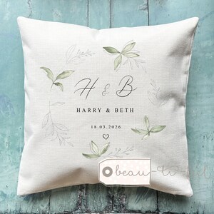 May include: Square, off-white decorative pillow with the initials "H & B" in elegant script, surrounded by a wreath of green leaves. The names "HARRY & BETH" and the date "18.03.2026" are below the initials, with a heart symbol.