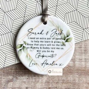 May include: White ceramic bauble with gold and green accents. The bauble features the text "Sarah & Jack I need an extra pair of hands to help me learn & grow. I know that yours will be the best as Mummy & Daddy told me so. Will you be my Godparents? Love, Amelia x"