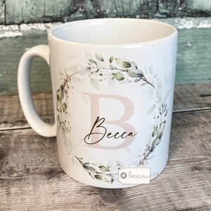 May include: White ceramic mug featuring a floral wreath design with the name "Becca" in a pink script font. The wreath is made of watercolour-style green leaves and pink flowers.