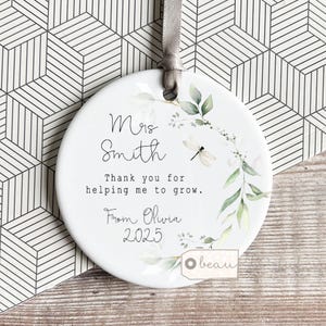 Può includere: Ornamento rotondo in ceramica bianca con un nastro grigio. Presenta la scritta "Mrs Smith" e "Thank you for helping me to grow." Illustrazioni floreali e di libellule adornano i bordi. Il nome "Olivia" e l'anno "2025" sono presenti.