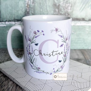 May include: White ceramic mug featuring a lavender floral wreath design with butterflies and the name "Christine" in script. The letter "C" is in a light purple color. The mug has a white handle and is sitting on a patterned surface.