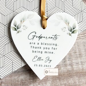 Könnte beinhalten: Weißer herzförmiger Keramik-Anhänger mit goldener Schleife. Der Anhänger zeigt ein Aquarell-Design mit grünen Blättern und dem Text "Godparents are a blessing. Thank you for being mine. Ellie Jay 25.03.2023".