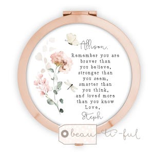 May include: A rose gold compact mirror with a floral design and inspirational text. The mirror features a watercolor illustration of pink flowers, a butterfly, and a dragonfly. The text reads "Remember you are braver than you believe..."