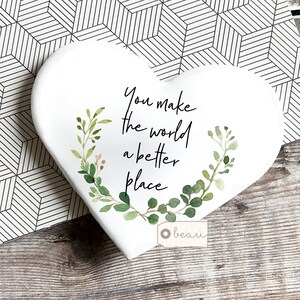 May include: White heart-shaped decorative piece with the text "You make the world a better place" in black script. Green watercolor leaves and sprigs surround the text. A small tag with the word "beau" is visible.