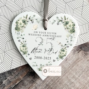 Puede incluir: Placa de cerámica en forma de corazón con un diseño floral y el texto "ON YOUR SILVER WEDDING ANNIVERSARY 25th Mum & Dad 2000-2025." La placa tiene una cinta gris para colgar y está decorada con mariposas y corazones.