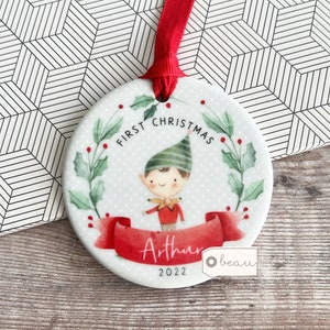 Könnte beinhalten: Weißer Keramik-Weihnachtsbaumschmuck mit einer Illustration eines grünen und roten Elfen, einem roten Bandbanner mit dem Text "First Christmas Arthur 2022" und einem grünen Stechpalmenkranz mit roten Beeren.