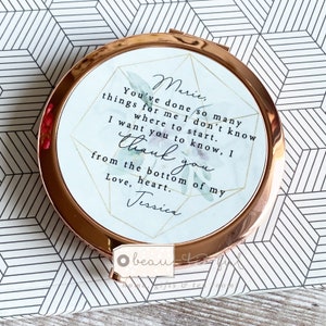 May include: A rose gold compact mirror with a white background and a floral design. The mirror has a message that reads "Marie, You've done so many things for me I don't know where to start. I want you to know, I thank you from the bottom of my heart. Love, Jessica"