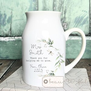 May include: White ceramic mug with a curved handle, decorated with a floral design of green leaves and white flowers. The mug has text that reads "Mrs. Smith, Thank you for helping me to grow. From Olivia 2025."