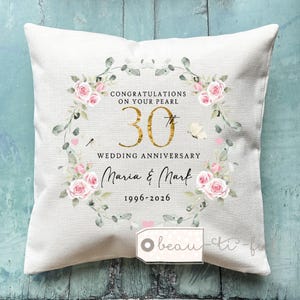 Op de afbeelding: Een vierkant, crèmekleurig kussen met een bloemenkransontwerp. De krans omcirkelt de tekst "Congratulations on your Pearl 30th Wedding Anniversary Maria & Mark 1996-2026". Het ontwerp bevat roze rozen en groene bladeren.