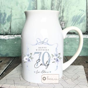 Puede incluir: Jarra de cerámica blanca con asa curva, decorada con un diseño floral azul y el texto "Happy Birthday 70 Emily Love Helen x". La jarra tiene una boca ancha y es adecuada para servir bebidas.