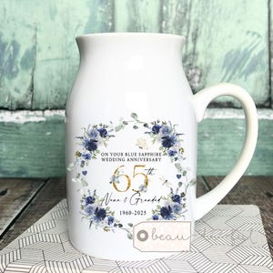 May include: White ceramic pitcher with a handle, decorated with a blue floral wreath and text. The text reads "ON YOUR BLUE SAPPHIRE WEDDING ANNIVERSARY", "65th", "Nana & Grandad", and "1960-2025". A small tag with the word "beau" is attached.