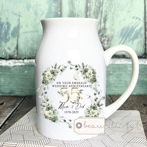 May include: White ceramic jug with a handle, decorated with a floral wreath and the text "ON YOUR EMERALD WEDDING ANNIVERSARY 55th Mum & Dad 1970-2025". The design incorporates gold detailing and green and white flowers.