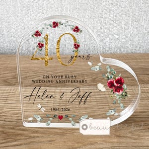 Può includere: Una targa in acrilico trasparente a forma di cuore con il numero 40 in oro, rose rosse e foglie di eucalipto. Include il testo ON YOUR RUBY WEDDING ANNIVERSARY, i nomi Helen & Jeff e gli anni 1986-2026 con un piccolo cuore rosso.