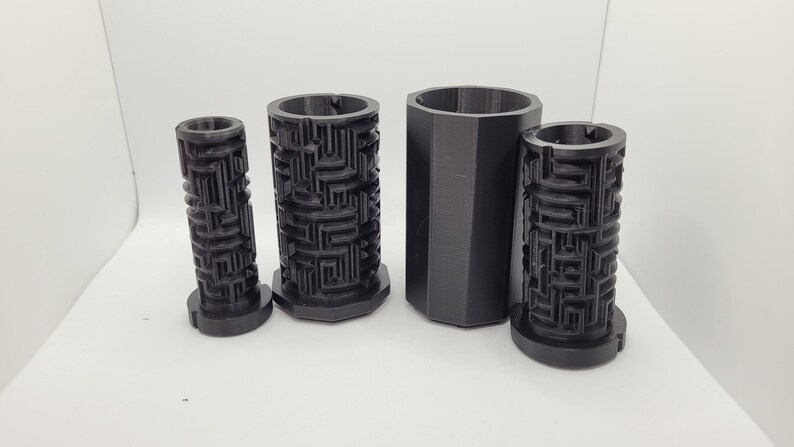 Op de afbeelding: Vier zwarte, 3D-geprinte cilindrische containers met een labyrintpatroon. De containers zijn van verschillende groottes en zijn in een rij gerangschikt van de kleinste tot de grootste.