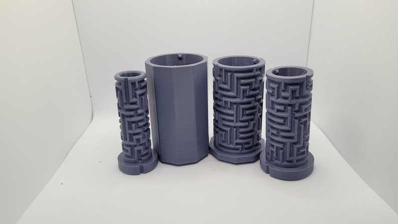 Op de afbeelding: Vier grijze 3D-geprinte cilinders met een labyrintpatroon. De cilinders zijn van verschillende grootte en zijn in een rij op een wit oppervlak gerangschikt.