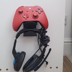 May include: A red video game controller and black headphones are mounted on a white wall with a black stand.