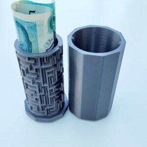 Op de afbeelding: Een grijze 3D-geprinte gelddoos met een labyrintpatroon aan de zijkant. Een opgerold biljet wordt in de doos gestoken. De doos is open en de andere helft ligt ernaast.