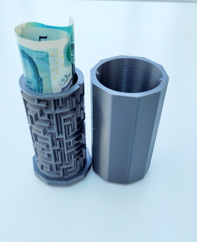 Op de afbeelding: Een grijze 3D-geprinte gelddoos met een labyrintpatroon aan de zijkant. Een opgerold biljet wordt in de doos gestoken. De doos is open en de andere helft ligt ernaast.