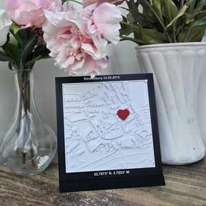 May include: A white 3D printed map of Shrewsbury, England with a red heart marking a location. The map is on a black background with the date "22.05.2015" and coordinates "52.7073° N, 2.7553° W" printed below the map.