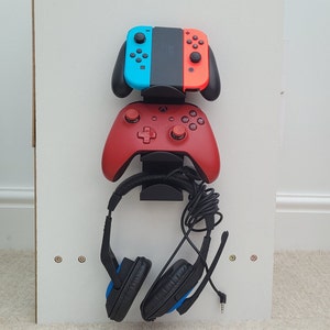 May include: A white wall mount with a black stand holding a red and black video game controller, a blue and black video game controller, and a black headset with blue earcups.