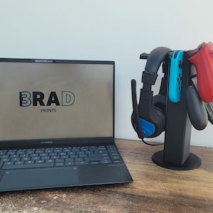 May include: A black stand with a blue headset, a blue and black Nintendo Switch controller, a red and black Xbox controller, and a black PlayStation controller. The stand is on a wooden surface next to a black laptop with the word "BRAD PRINTS" on the screen.