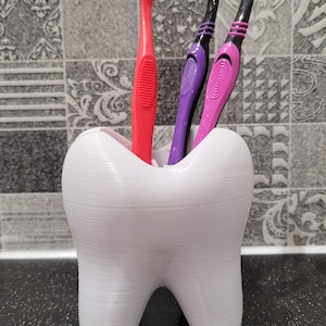 May include: A white tooth-shaped toothbrush holder with three toothbrushes inside. The toothbrushes are pink, purple, and black.