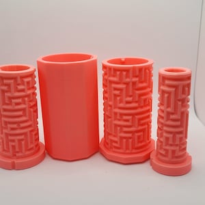 Op de afbeelding: Vier roze plastic mallen voor het maken van kaarsen. De mallen zijn cilindrisch en hebben een labyrintpatroon aan de buitenkant.
