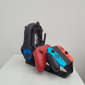 May include: A black gaming headset, a red Xbox controller, and a blue and orange Nintendo Switch controller are displayed on a black stand.