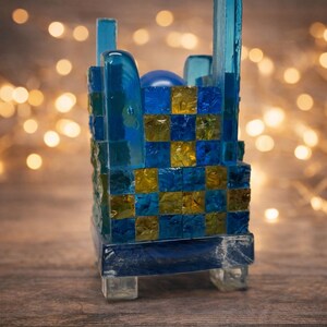Può includere: Un portacandele decorativo in vetro con un design a mosaico. Il supporto presenta un mix di quadrati blu e oro, con una sfera blu all'interno. Il supporto è sostenuto da piedini in vetro trasparente.