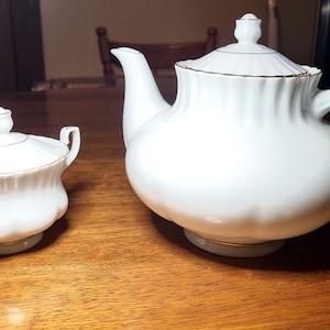 Op de afbeelding: Een wit porseleinen theeservies, bestaande uit een theepot met deksel en een suikerpot met deksel. Beide stukken hebben gouden randen en staan op een houten tafel. De theepot heeft een gebogen handvat en tuit.