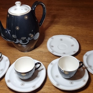 Puede incluir: Juego de té vintage en blanco y negro con detalles dorados. El juego incluye una tetera, tres tazas y platillos. La tetera y las tazas tienen un diseño de estrella. El juego está sobre una superficie de madera.