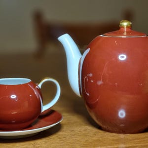 Puede incluir: Un juego de té rojo y blanco con detalles dorados. El juego incluye una tetera, una jarra de crema y un platillo. La tetera tiene forma redondeada con boquilla y asa blancas. La jarra de crema es pequeña y redonda con un diseño a juego.