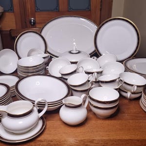 Puede incluir: Un juego completo de vajilla de porcelana blanca con ribete marrón oscuro y dorado. El juego incluye platos, cuencos, tazas, platillos, una fuente, una fuente cubierta y una jarra de crema.