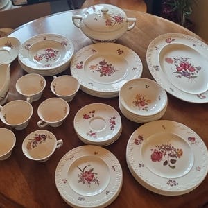 Op de afbeelding: Een verzameling antiek wit porseleinen serviesgoed met een bloemmotief. De set bevat borden, schotels, kopjes, een juskom en een serveerschaal met deksel. Het bloemmotief heeft rode, roze en gele bloemen.