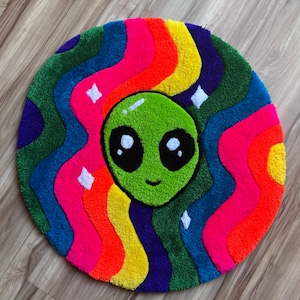 May include: A round, colorful rug with a green alien face in the center. The rug has a rainbow swirl pattern with shades of pink, orange, yellow, green, blue, and purple.